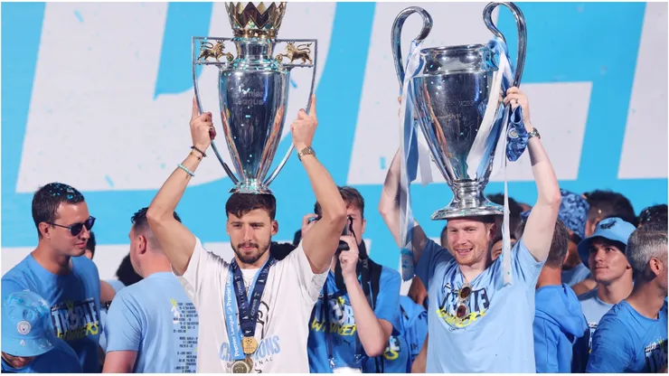 Ruben Dias of Manchester City celebrates with the Premier League Trophy as Kevin De Bruyne celebrates with the UEFA Champions League Trophy