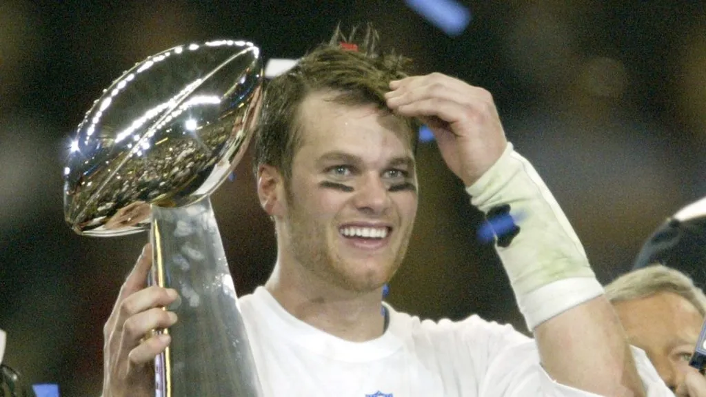 Tom Brady #12 of the New England Patriots raises the Lombardi trophy as he is interviewed by television commentator Jim Nance after defeating the Carolina Panthers 32-29 in Super Bowl XXXVIII at Reliant Stadium on February 1, 2004 in Houston, Texas.