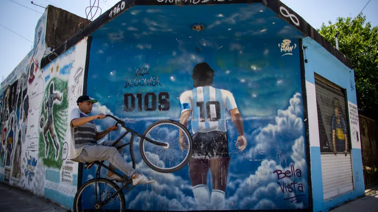BUENOS AIRES, ARGENTINA - OCTOBER 21: A man wheelies past a mural depicting Diego Armando Maradona entering heaven with a legend that reads in spanish "Here rests god" making reference to the fact that the idol is buried one block away from the mural in Bella Vista on October 21, 2021 in Buenos Aires, Argentina. Football fans all over the country remember their late idol, who would have turned 61 on October 30. (Photo by Tomas Cuesta/Getty Images)
