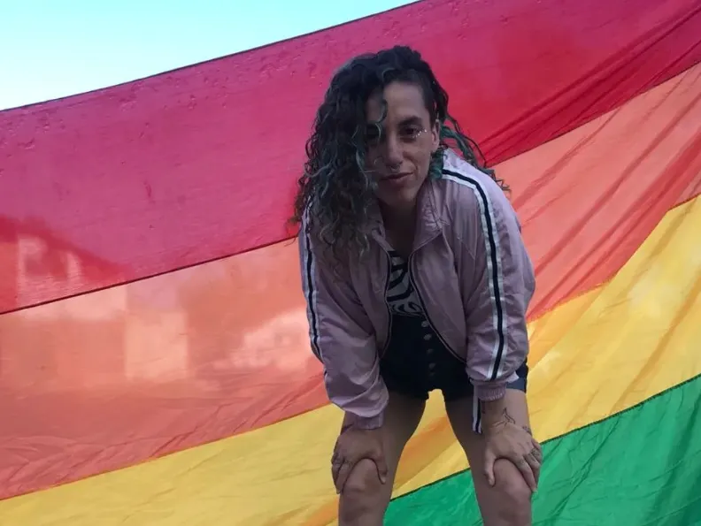 Maquinita Orellana posando junto a la bandera del orgullo, o bandera LGTB