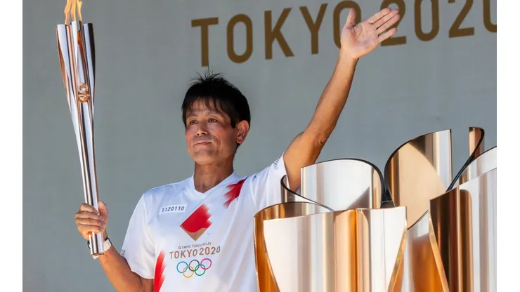 Um dos carregadores da tocha olímpica posa empunhando-a e acenando durante evento em Tóquio (Foto: Getty Images)