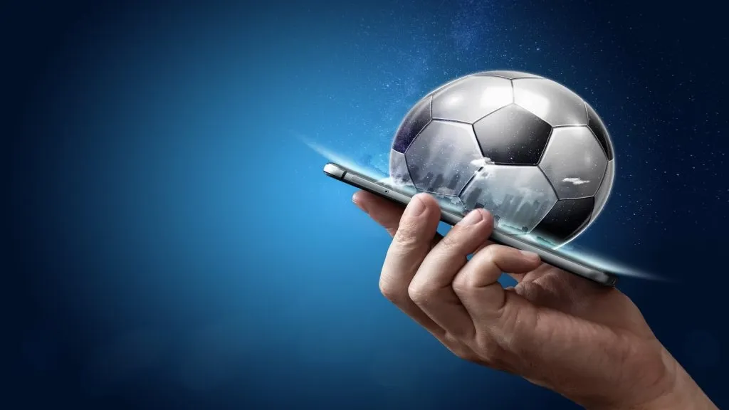Imagem gráfica de bola de futebol saindo de smartphone contra fundo azul. 