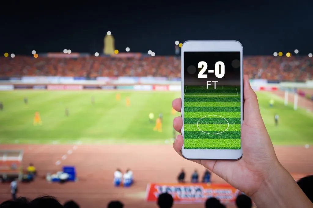 Smartphone sustendido por mão contra fundo de partida de futebol em estádio. Na tela, pode-se ler 