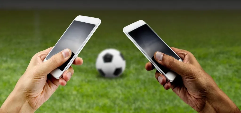 Foto das mãos de duas pessoas distintas segurando dois smartphones, uma branca à esquerda e uma negra à direita. Ao fundo, uma bola de futebol pousando em um gramado verde, simbolizando pessoas que apostam nas novas casas de apostas do Brasil. 