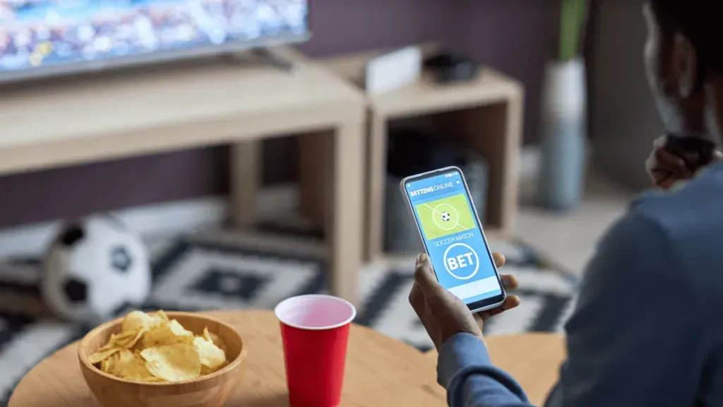 Imagem de mesa de madeira com vaso com chips e copo de plástico vermelho, bola de futebol e TV ao fundo, e imagem parcial de uma pessoa apostando ao vivo no celular