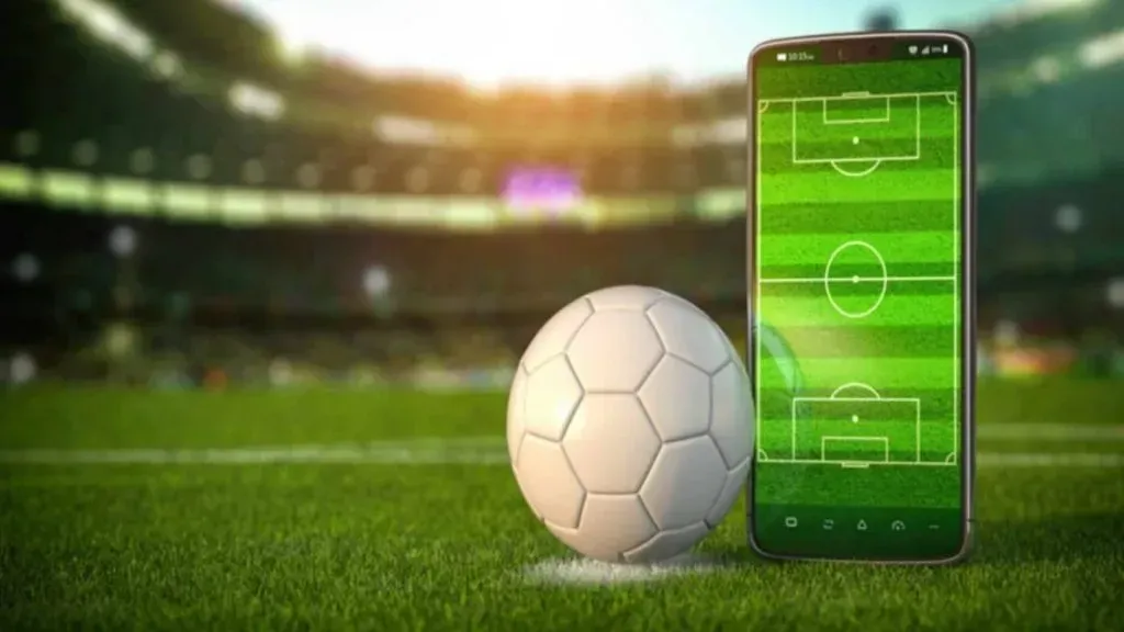 Uma bola branca de futebol e um celular com a tela representando um campo de futebol estão sobre o gramado do estádio