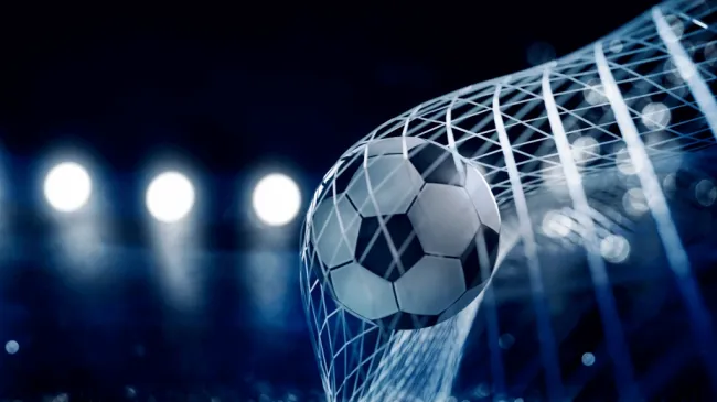 Bola de futebol clássica, em branco e preto, atingindo o fundo da rede, com holofotes ao fundo. 