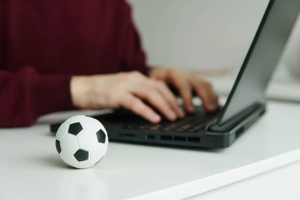 Pessoa usando notebook, com as duas mãos em cima do teclado, com uma mini bola de futebol clássica, em preto e branco, posicionada do seu lado direito. 