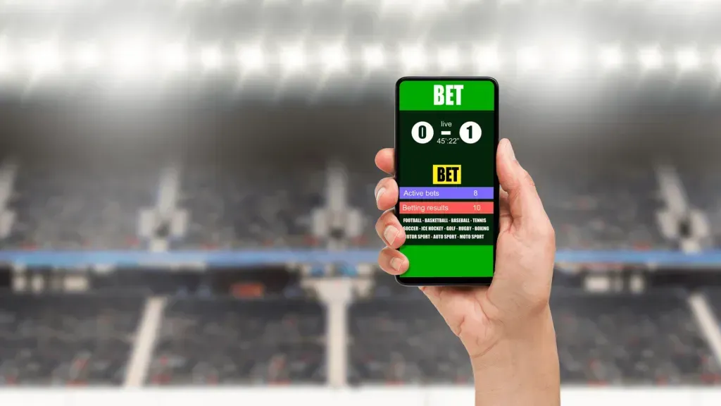 Uma mão segura um celular onde se vê o placar 0-1 de uma partida ao vivo, e de fundo uma arquibancada de estádio.