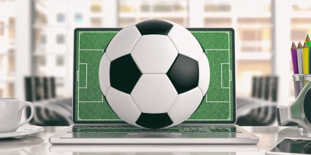 Imagem de notebook com bola de futebol clássica em cima do teclado. Ao lado esquerdo, xícara de café, do direito, porta-lápis com lápis de várias cores.