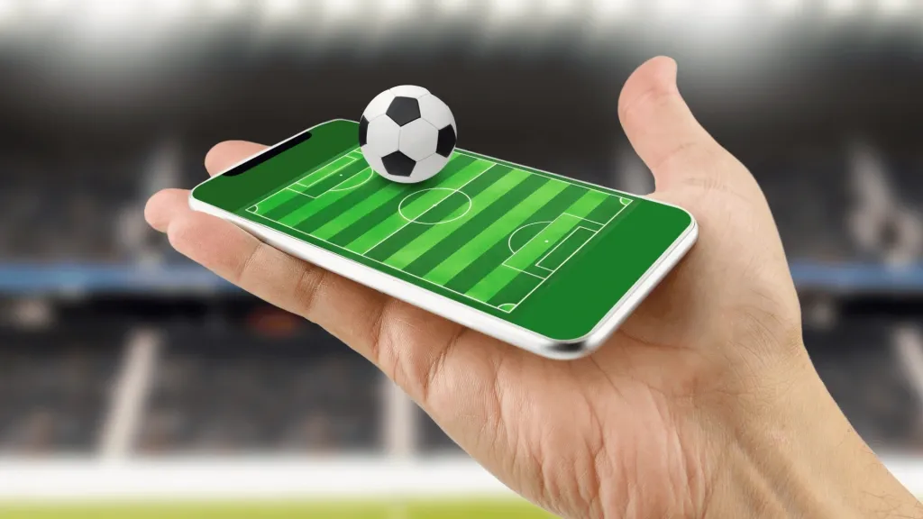 Mão segurando smartphone com campo de futebol projetado na tela. Na superfície, bola de futebol clássica em 3d, em tons de branco e preto.