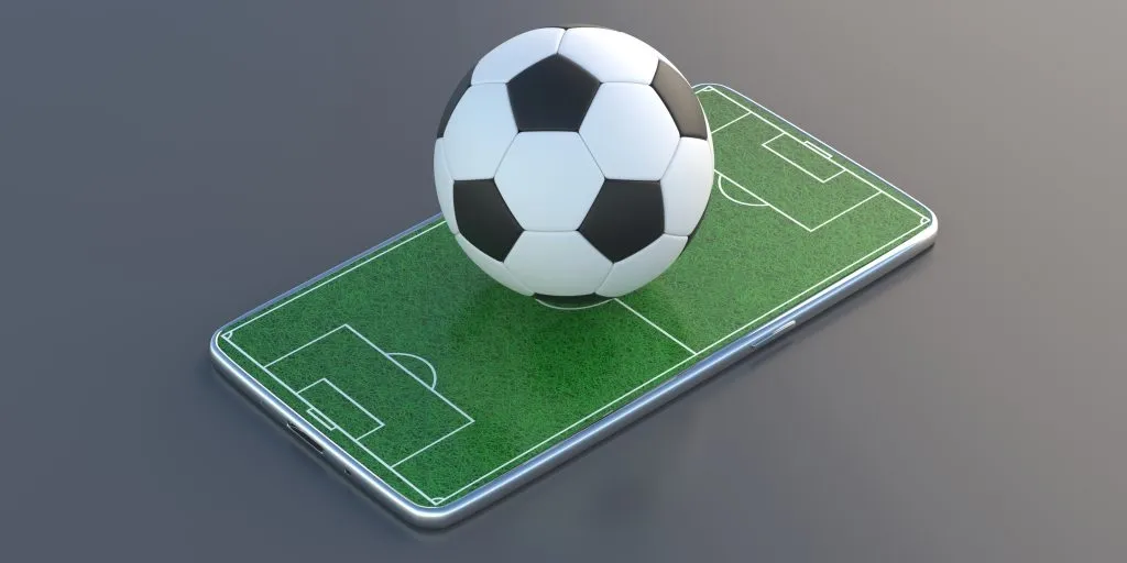 Smartphone com projeção de campo de futebol na tela. Na superfície, bola de futebol clássica, em branco e preto, em miniatura. 
