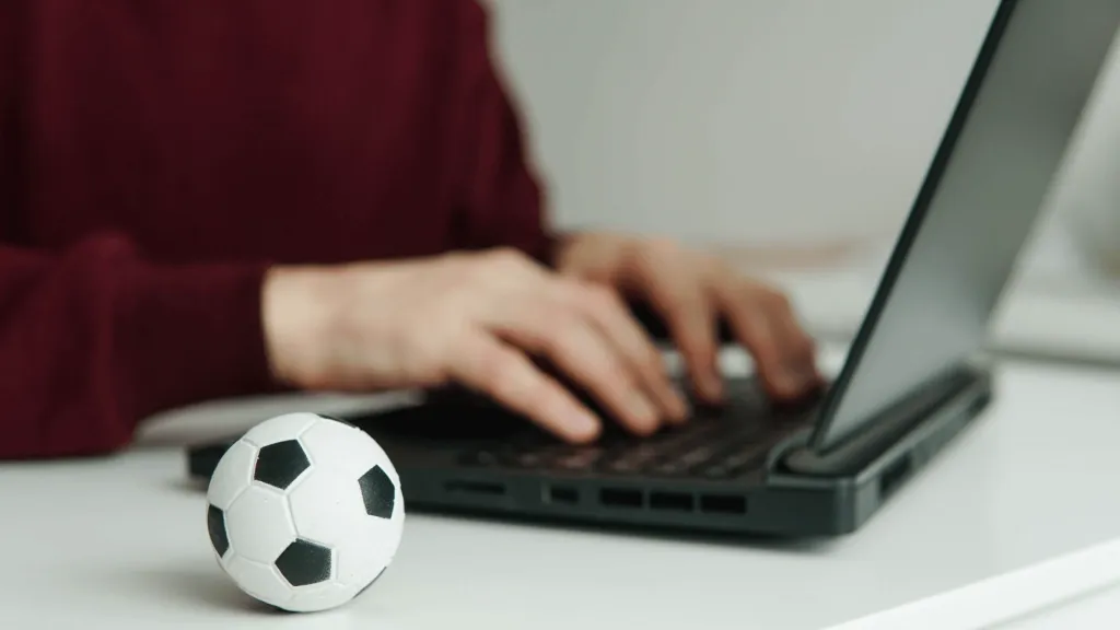 Foto de pessoa em notebook, usando suéter vermelho, com miniatura de bola de futebol clássica, em branco e preto, ao lado, simbolizando as apostas na Estrela Bet. 