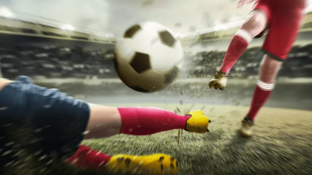 Imagem de dois jogadores, em bola dividida, o da esquerda ao chão, no gramado, em posição de carrinho, enquanto o outro tenta alcançar a bola com um chute. 